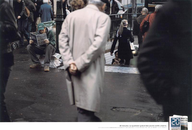 Publicité 20 minutes - 20 minutes. Le quotidien des jeunes actifs urbains - Le distributeur de journaux Métro est assis sur sa pile de journaux pour lire le concurrent. - Agence BDDP&Fils  