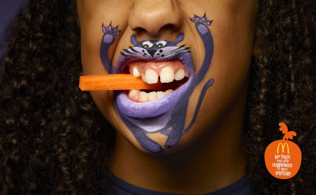 Publicité McDonald's - We teach your little carnivorous to enjoy fruit/Vegetable - Fillette avec un maquillage de panthère autour de la bouche a un batonnet de carotte entre ses dents - Agence TBWA - Espagne - 2018